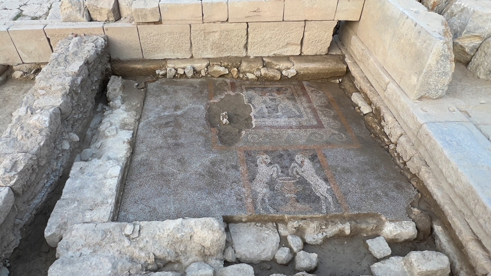 An excavation site. In the center are two mosaics on brown dirt, surrounded by a low rock wall and then higher limestone walls on three sides. 